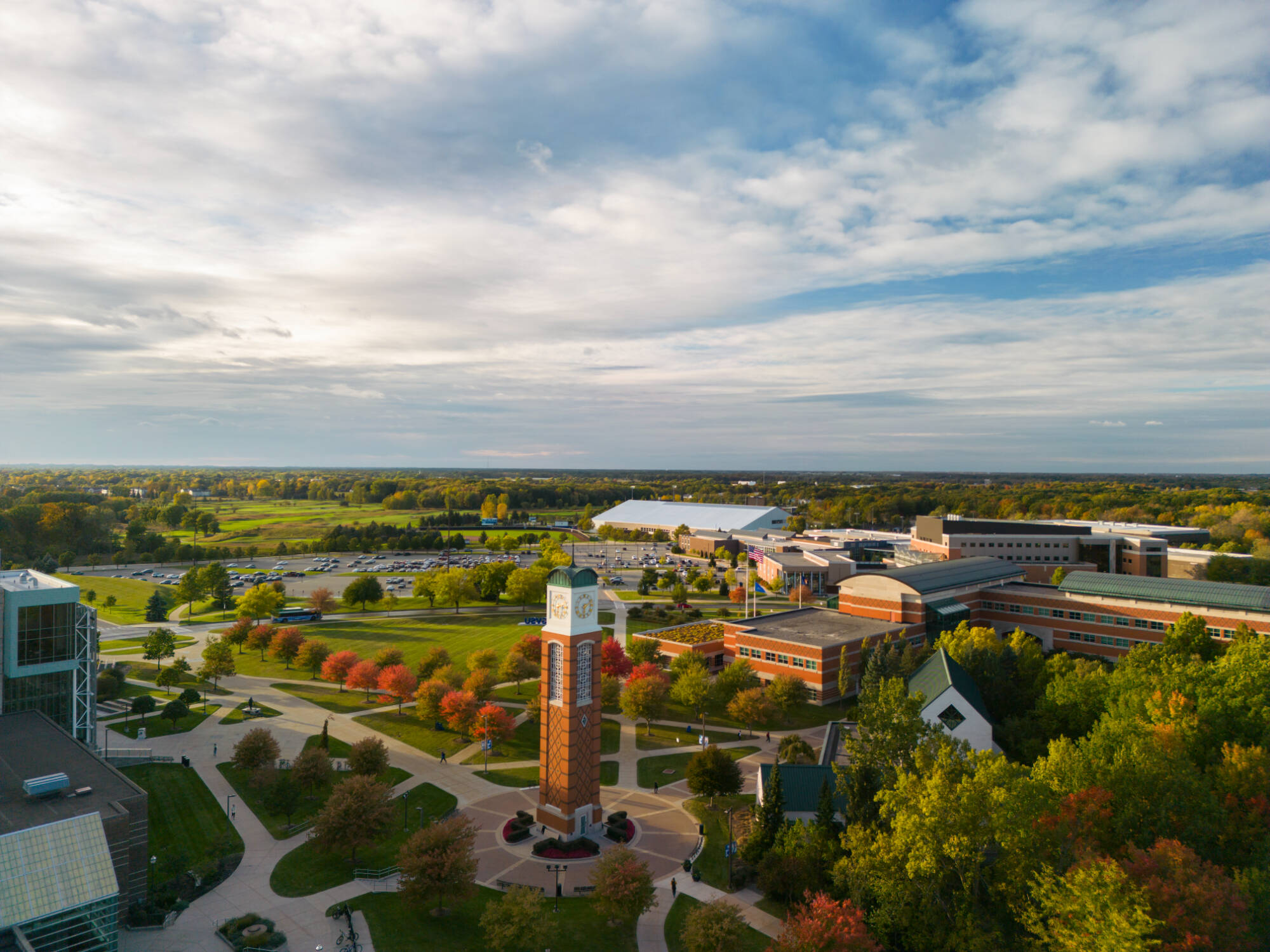 An aerial view of a university campus with a prominent clock tower, colorful autumn trees, and modern buildings under a partly cloudy sky.